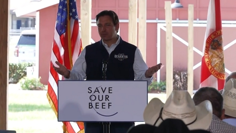 Governor speaking at podium with Save Our Beef sign and US flags behind